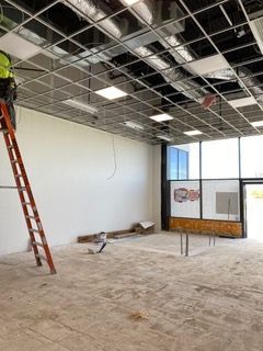 Person on a ladder installing ceiling tiles in an empty room with large windows, concrete floor, and white walls.