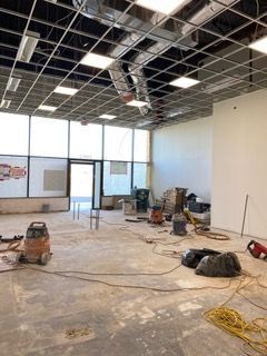 Interior space under construction; exposed ceiling grid, concrete floor, equipment and materials scattered.