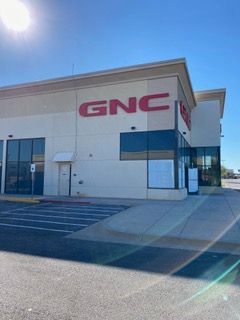 GNC store exterior with large red logo on a cream-colored building. Clear blue sky, sunny day.