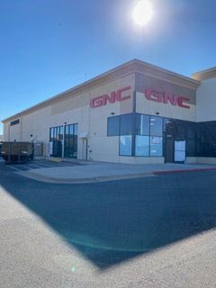 GNC store exterior with large red logo on a sunny day. Beige building with storefront windows and a dark shadow.