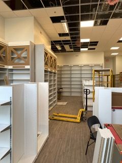 Interior of a store under construction with empty shelves, a yellow pallet jack, and exposed ceiling tiles.