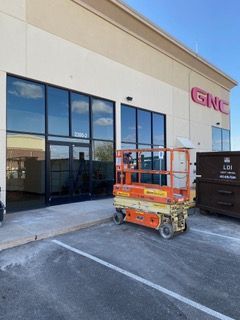 GNC store exterior with an orange lift in front of the window and a dumpster on the side.