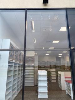 View through a storefront window of an interior retail space with white shelving and bright lighting.