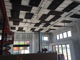 Interior construction site with black acoustic panels installed on the ceiling. A worker in a lift is visible.