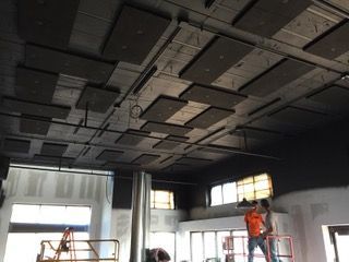 Workers installing sound-dampening panels on a dark-painted ceiling in a building under construction.