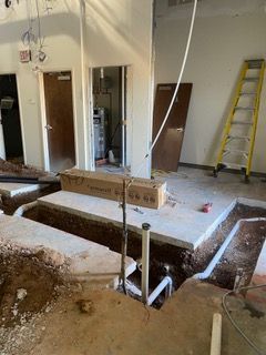 Construction site with trenches dug in the floor, pipes, concrete, and doors in the background. A ladder leans against the wall.