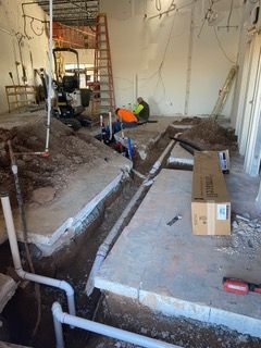 Construction site: Trench with pipes in concrete floor. Workers in orange vests are present.