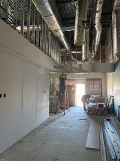 Interior under construction; workers on a ladder installing ductwork in a building. Metal studs, exposed ducts, and drywall visible.
