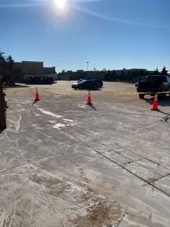 Sunlit parking lot with orange cones and police tape. Vehicles and a building are visible in the background.