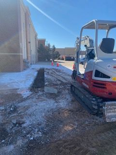 Mini excavator on a partially cleared pathway next to a building on a snowy, sunny day.