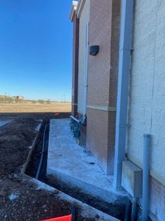 Construction site with trench and concrete against a building's brick and stucco facade, blue sky overhead.