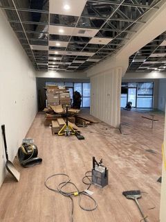 Interior under construction with exposed ceiling grid, wood flooring, and construction materials.