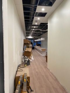 Interior retail space under construction: long hallway with unfinished ceiling, light-colored walls, wood-look flooring. Boxes and tools are present.