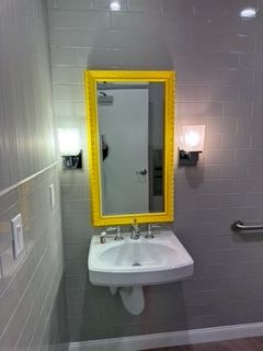 Yellow-framed mirror above a sink in a public restroom. Gray tile walls with sconce lighting.