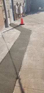 A section of freshly poured concrete sidewalk, next to a building with an orange traffic cone in the middle.