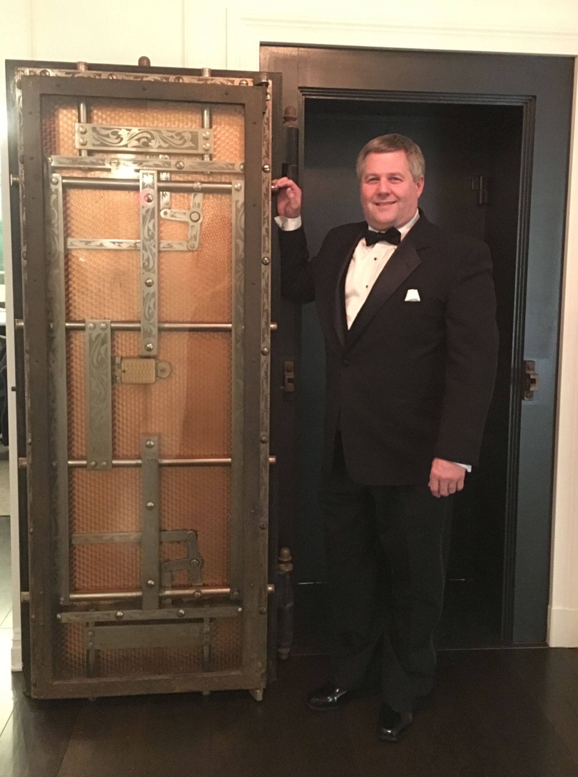 Man in tuxedo stands beside an ornate metal door in a room with dark doorway.