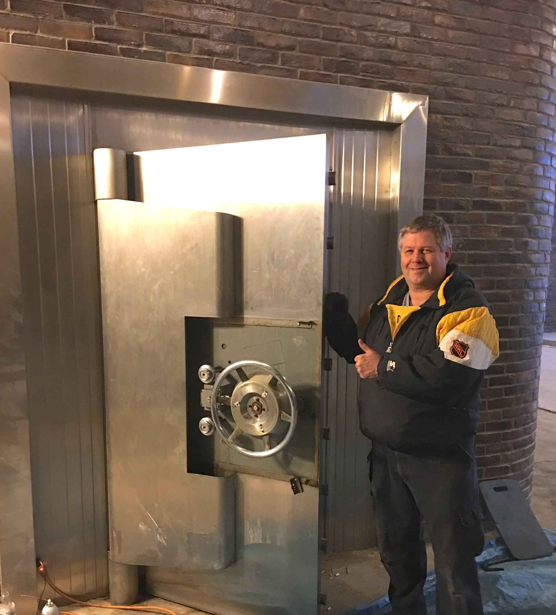 Man gives thumbs up beside large, open bank vault door in a brick building.