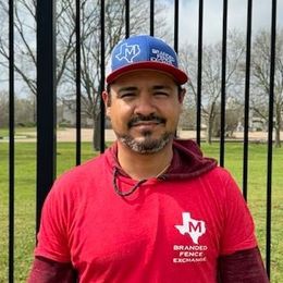 A man wearing a hat and a red shirt is standing in front of a fence.