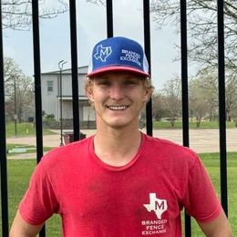 A man wearing a red shirt and a blue hat is standing in front of a fence.