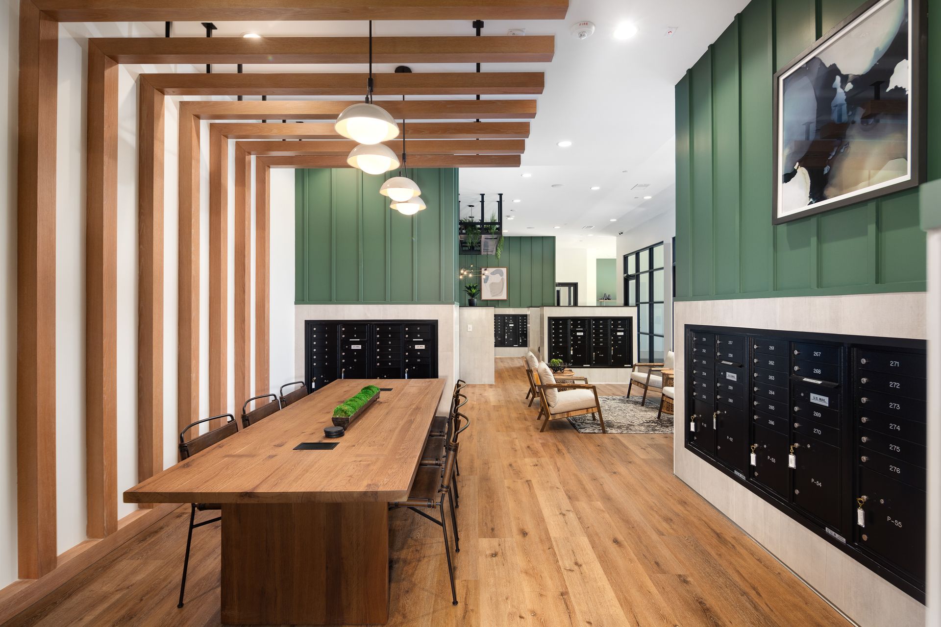 Modern office lounge with wooden tables, green walls, hanging lights, and black wall-mounted display boards