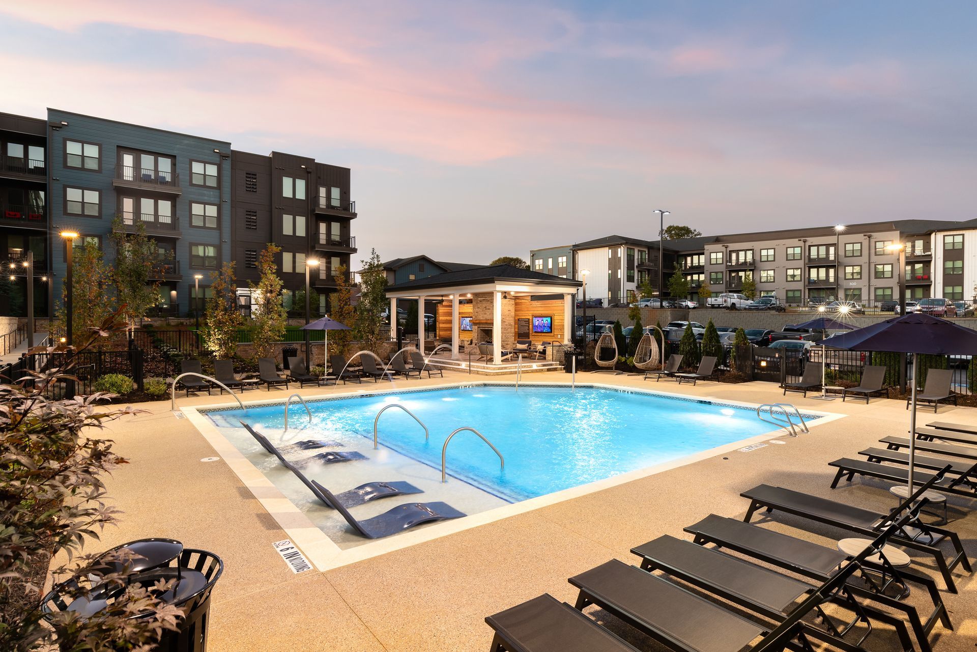 Apartment pool at dusk with lit water, lounge chairs, and surrounding buildings