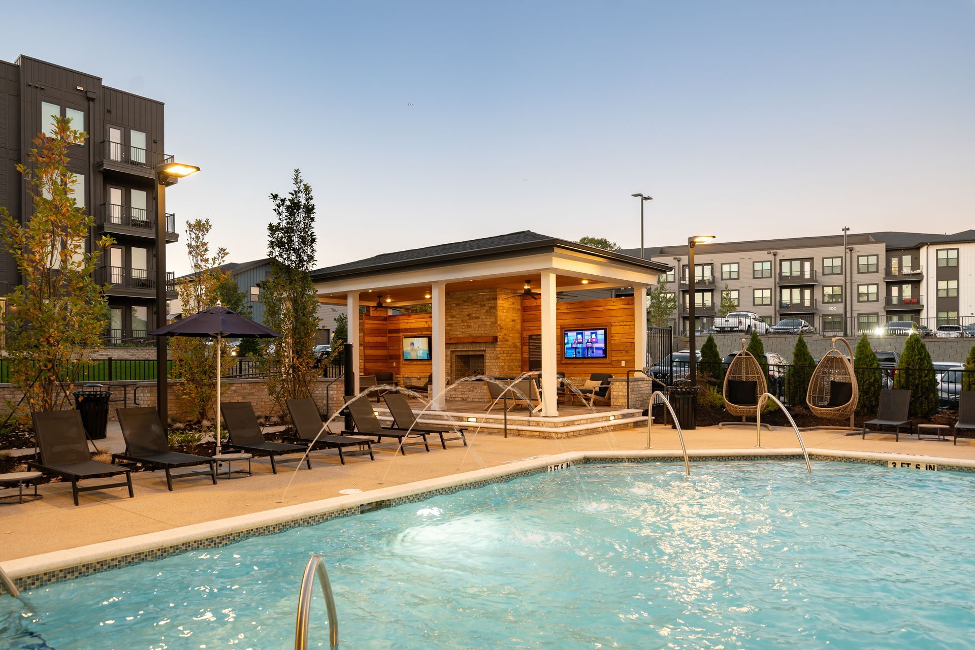 Apartment pool area at dusk with lounge chairs, cabana, and modern buildings nearby