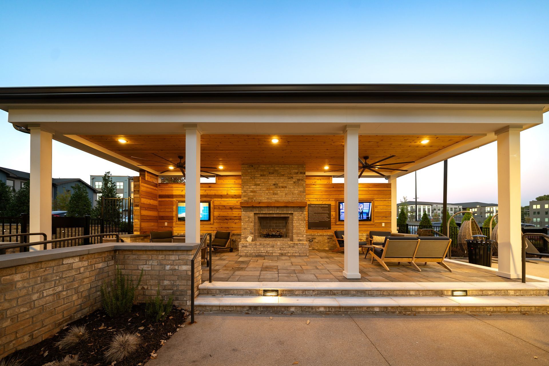 Covered patio with stone fireplace, wooden ceiling lights, and lounge chairs at dusk.