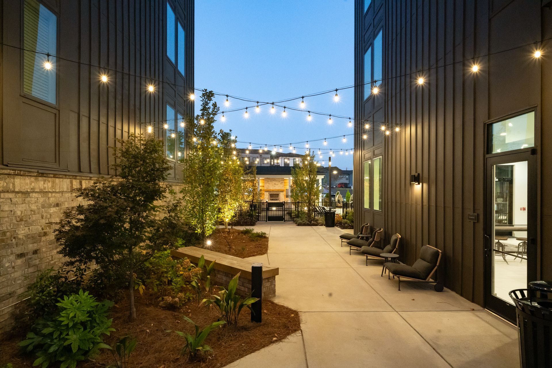 Courtyard walkway between modern buildings with string lights, seating, and plants at dusk