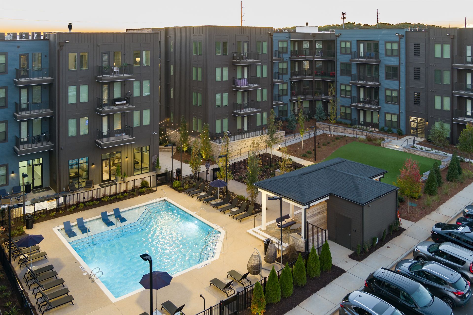 Apartment complex courtyard with swimming pool, lounge chairs, and parked cars at sunset
