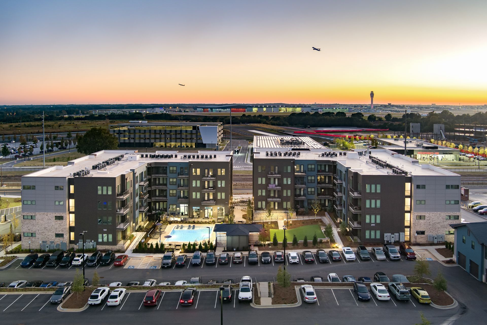 Aerial view of a modern apartment complex at sunset with parked cars and city lights in the distance