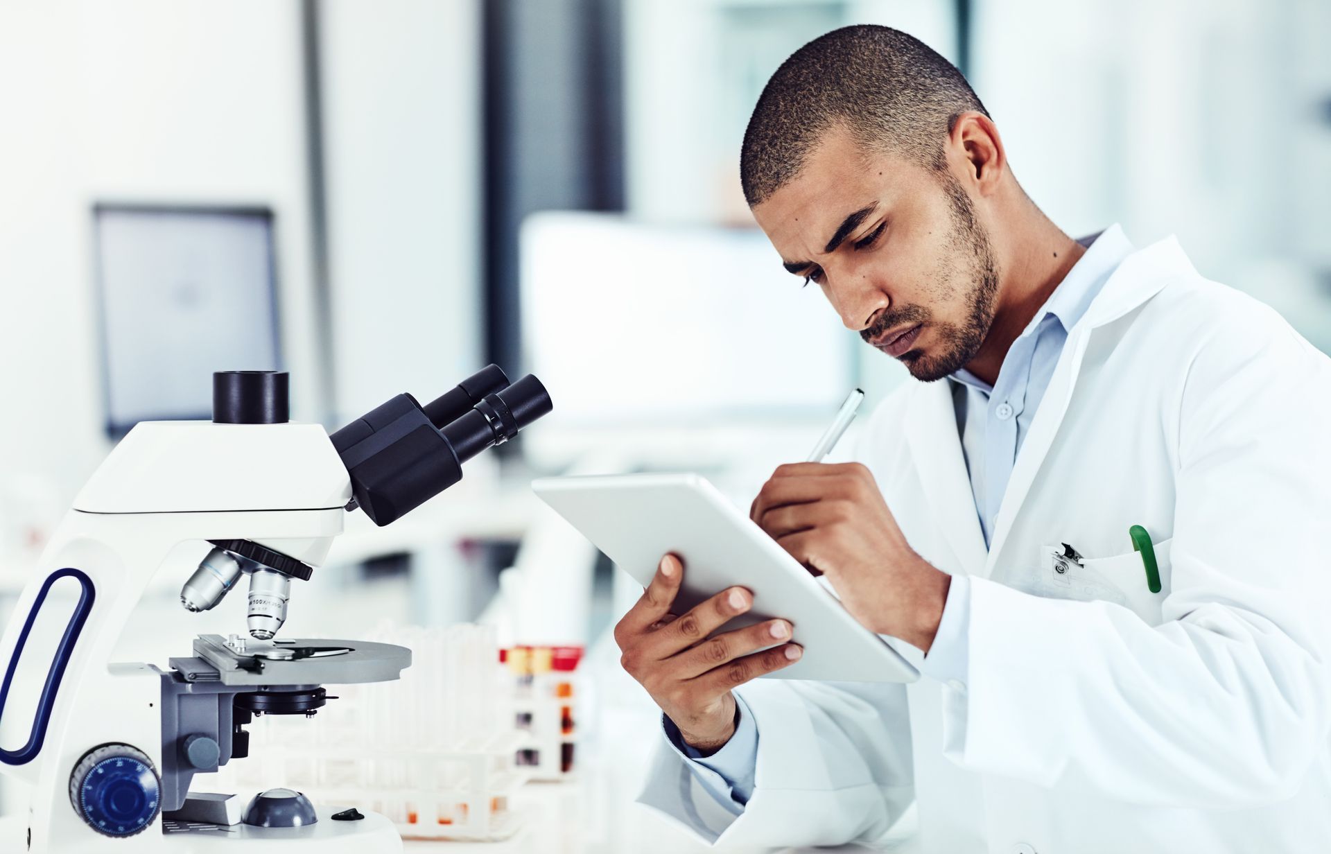 A man in a lab coat is looking at a tablet in front of a microscope.