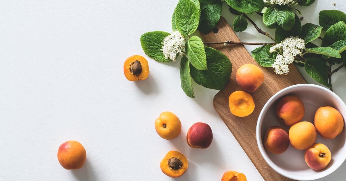 A bowl of apricots next to a cutting board and flowers on a table.