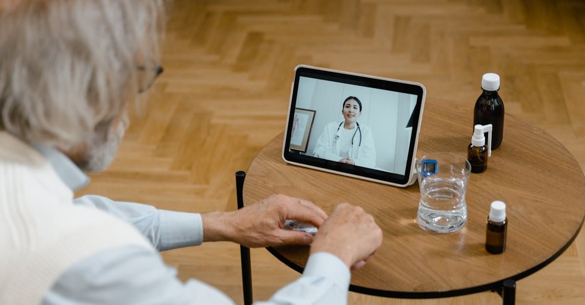 An elderly man is sitting at a table using a tablet computer to have a video call with a doctor.