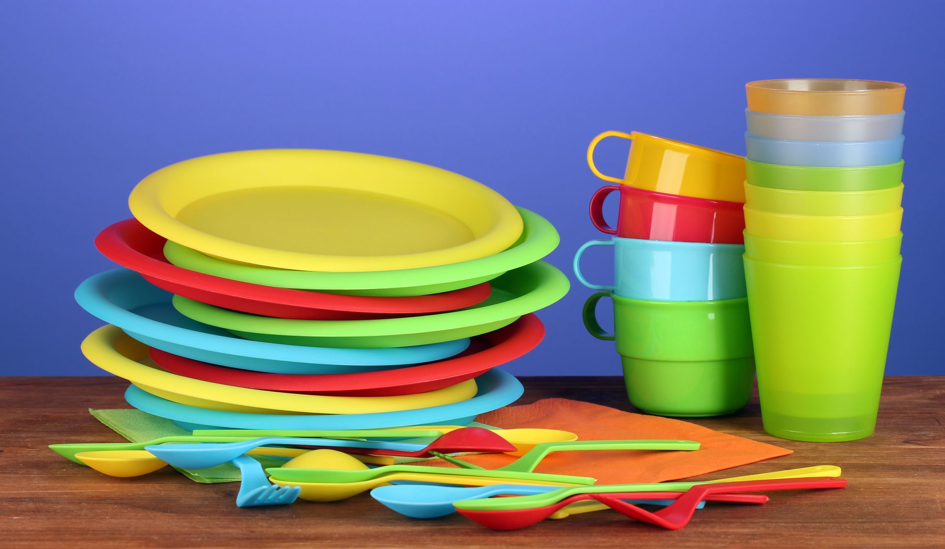 A stack of colorful plastic plates , cups , spoons and napkins on a wooden table.