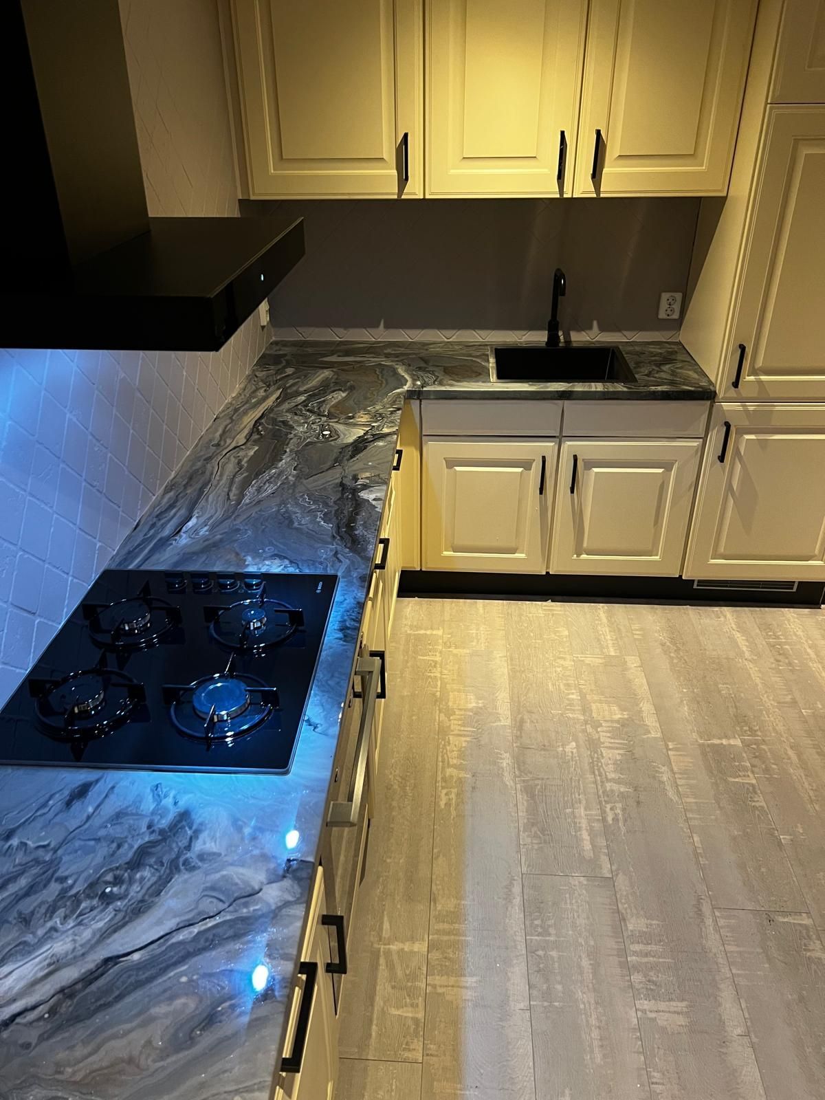 Keuken met crèmekleurige kasten, donkere aanrechtbladen, kookplaat en spoelbak.