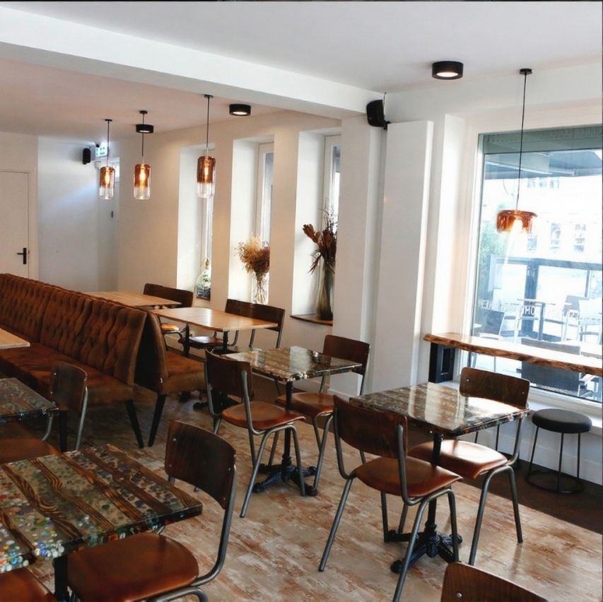 Interieur van een café met tafels, stoelen en een lange bank. Koperen hanglampen hangen aan het plafond.