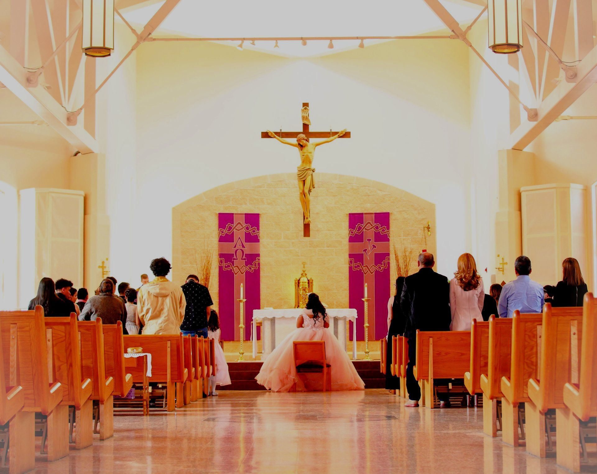 A woman in a pink dress sits on a bench in front of a cross in a church