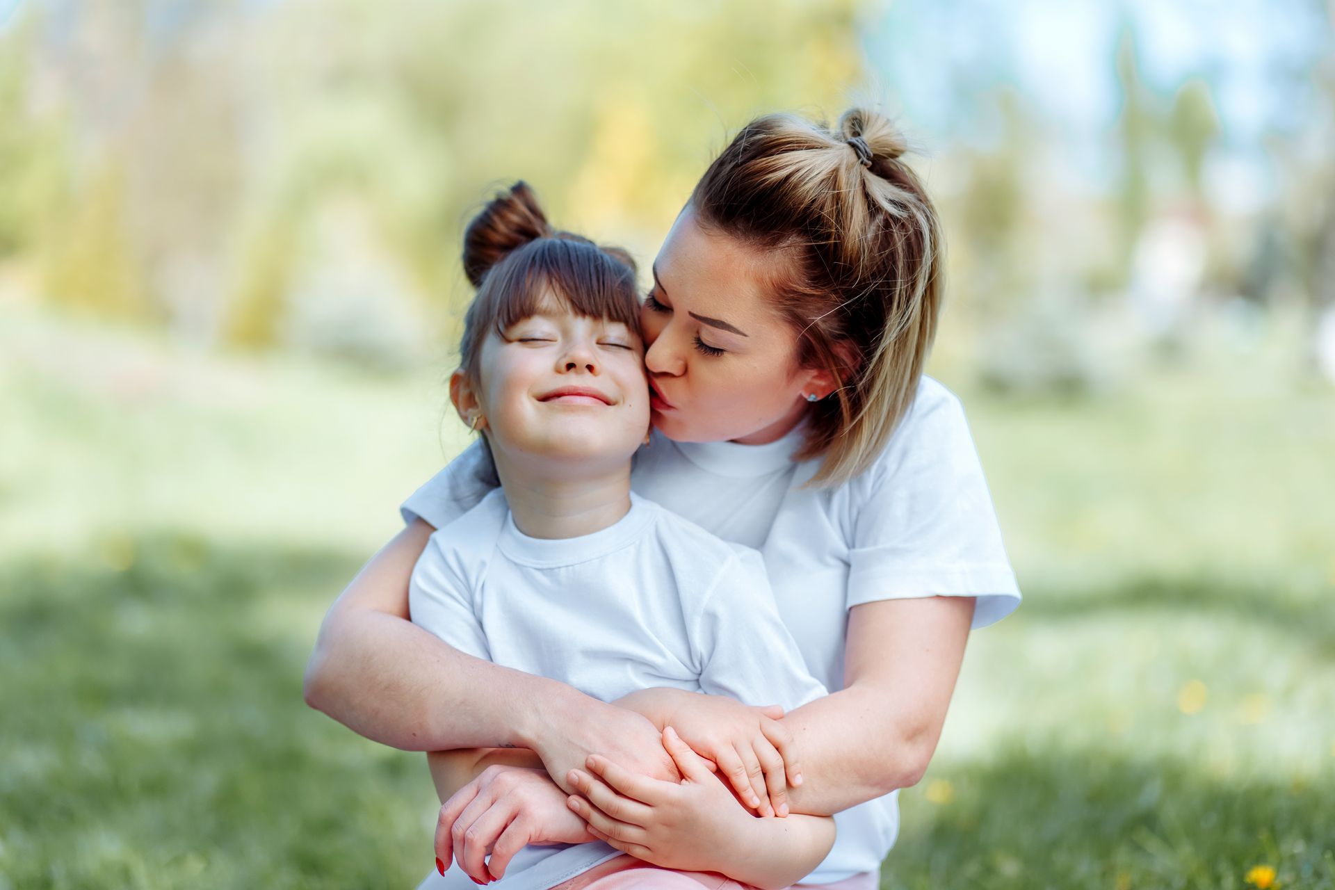 A woman is kissing her daughter on the cheek in a park.