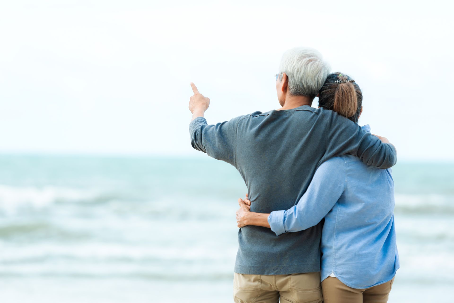 An elderly couple is standing on the beach looking at the ocean.