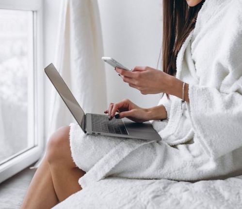 Uma mulher de roupão está sentada em uma cama usando um laptop e um celular.-Pousada-Casa-Flor