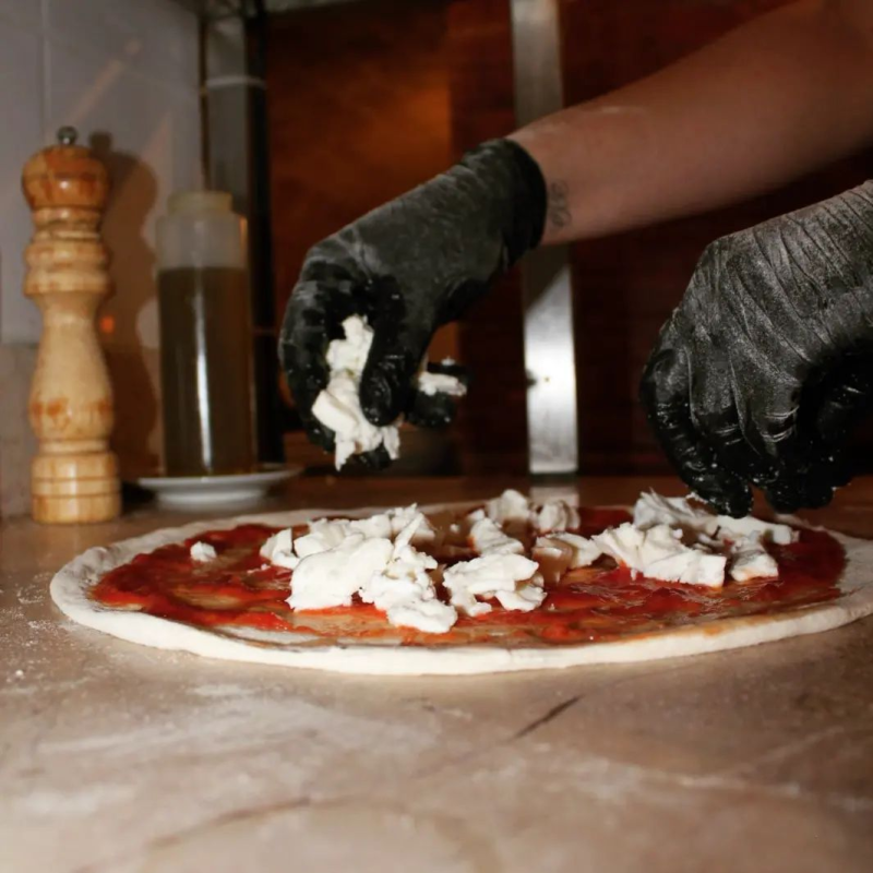 Una persona che indossa guanti neri sta mettendo il formaggio su una pizza