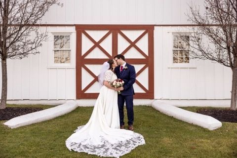 A bride and groom are kissing in front of a white barn.