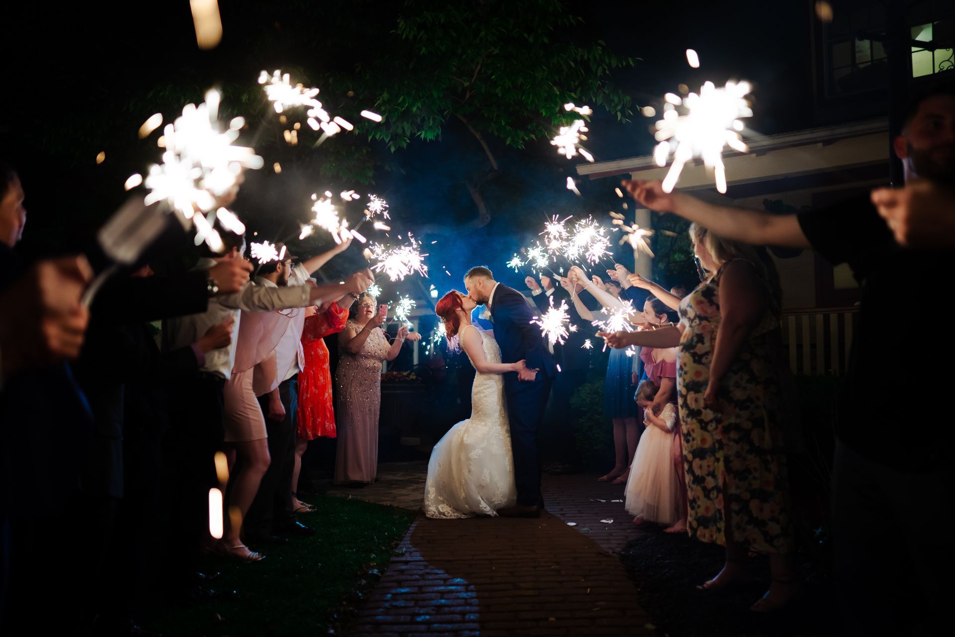 Newlyweds kiss as guests wave sparklers, night wedding celebration.
