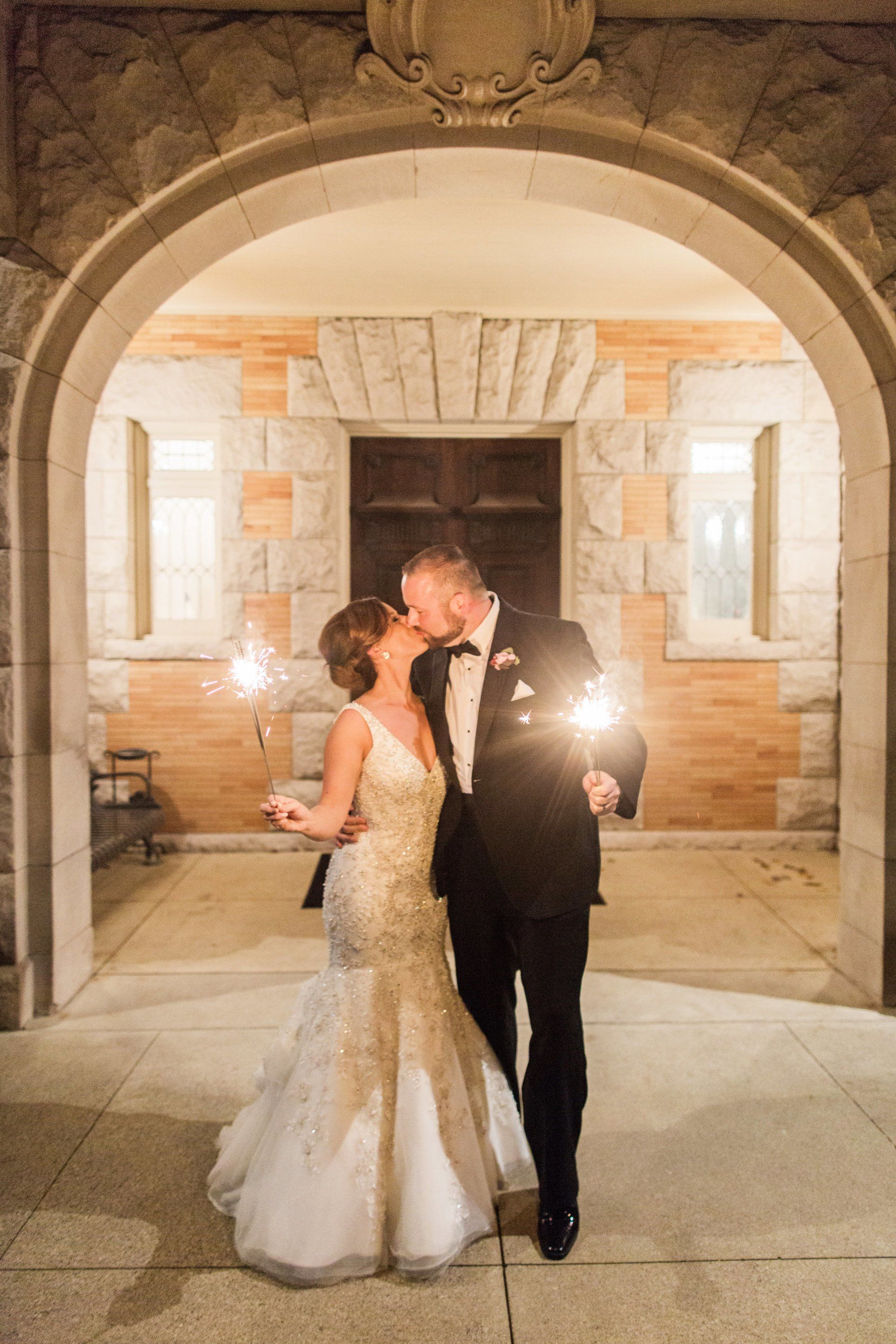 A bride and groom are kissing while holding sparklers in front of a building.
