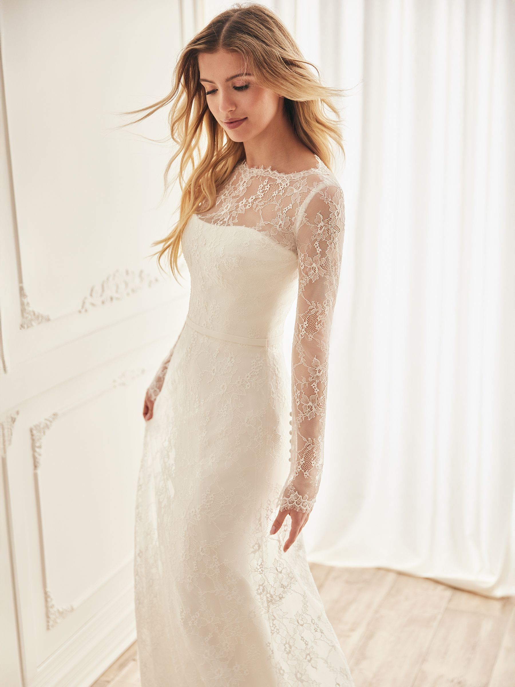 Woman in a white lace wedding dress with long sleeves, looking down, in a bright room.