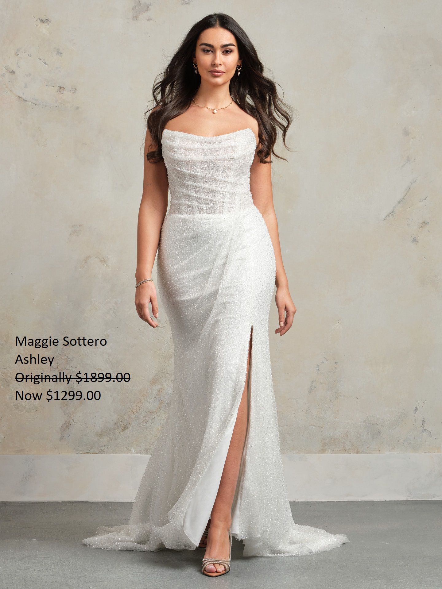 Woman in a white strapless gown with a high slit, posing in front of a neutral backdrop. Maggie Sottero Ashley