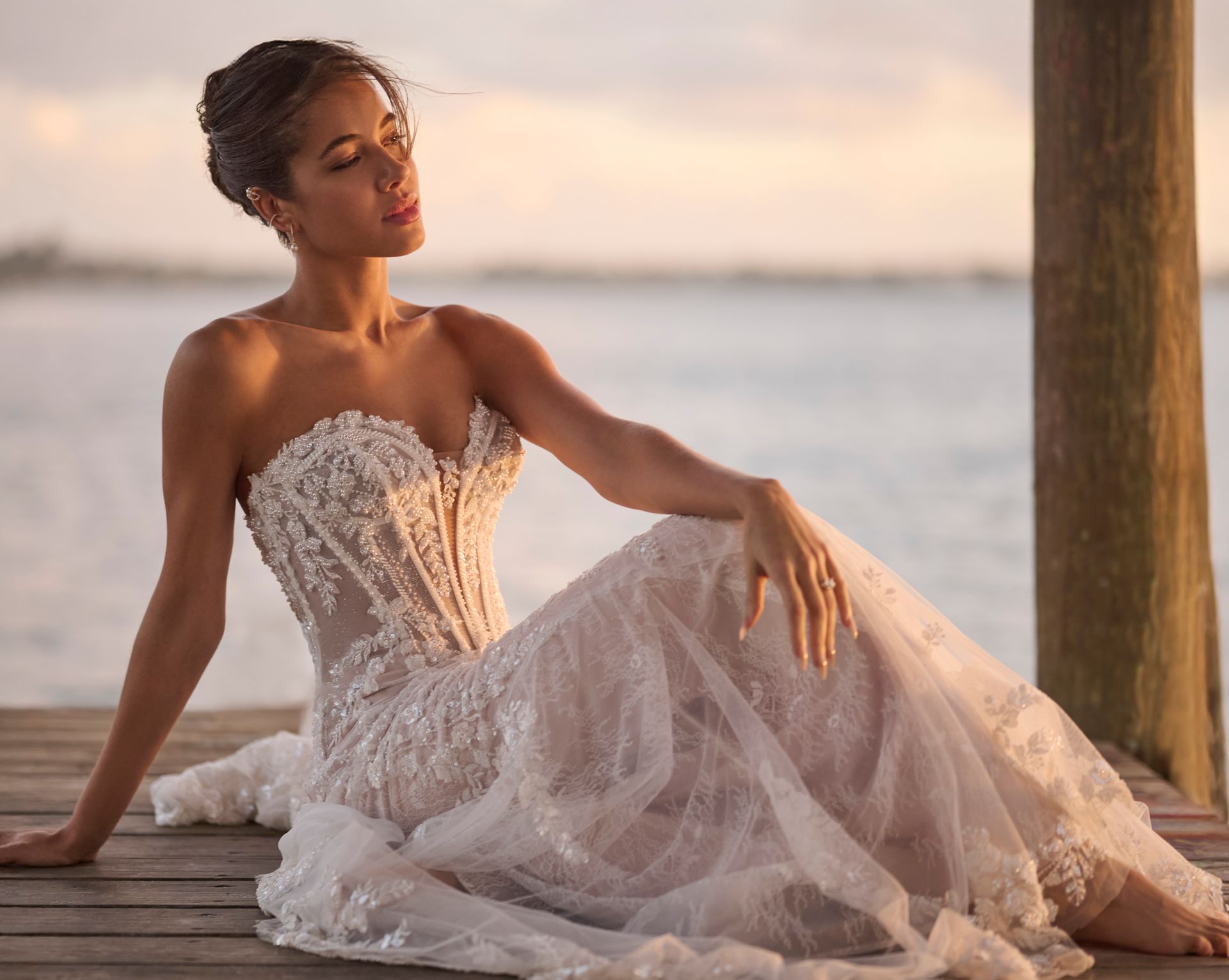 Woman in strapless wedding dress, seated on wooden dock, eyes closed, sunset backdrop.