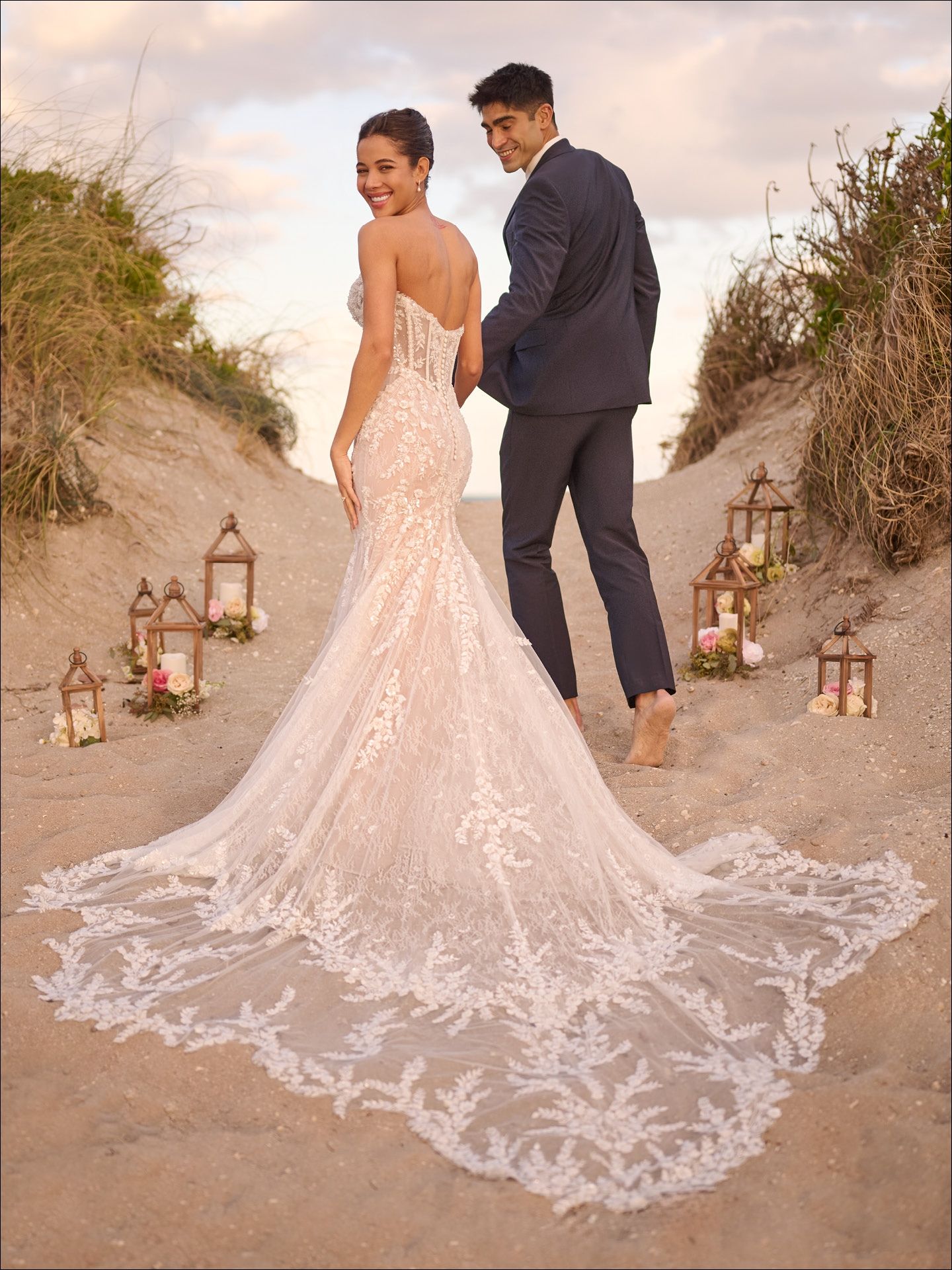 Bride and groom on sandy path. Bride in white lace dress, groom in suit. Beach setting, lanterns.