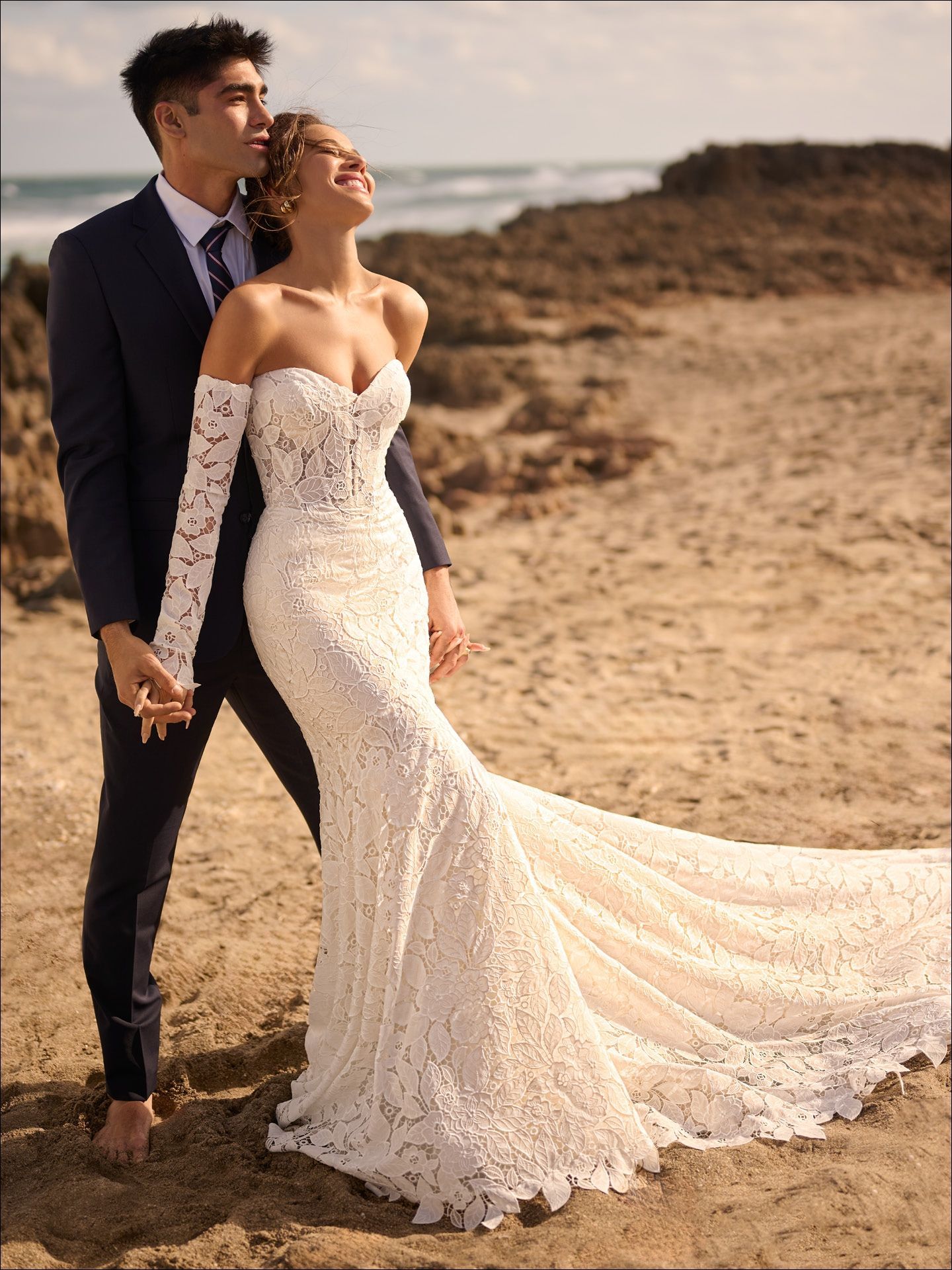 Couple on a beach: Woman in a white lace wedding dress, man in a suit, holding hands, ocean in background.