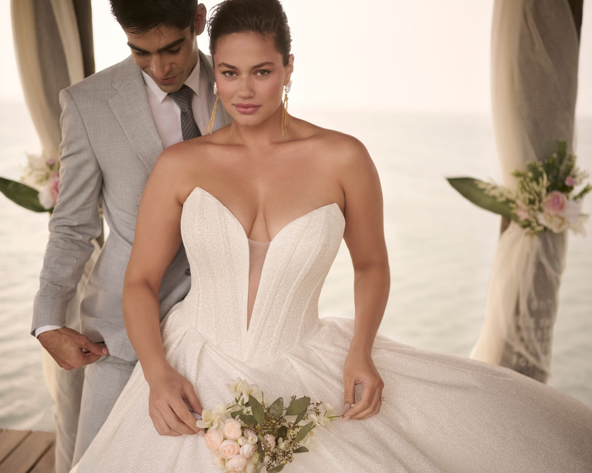Bride in sparkling white gown, groom in gray suit, posing outdoors.